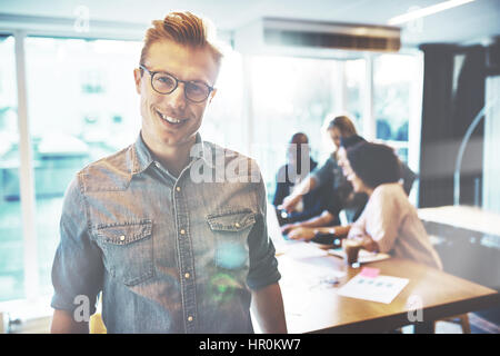 Giovane uomo bello indossare occhiali e abiti casual guardando la fotocamera e sorridente, stando in ufficio luminoso con il team lavora in background Foto Stock