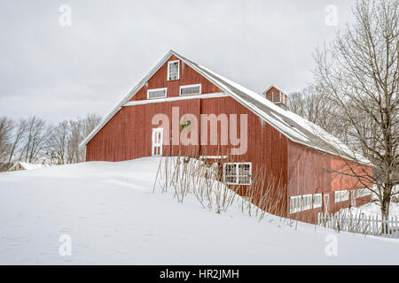 Un tradizionale in legno rosso incorniciato cavallo fienile in inverno con neve sul terreno e una ghirlanda di Natale sulla porta a Enfield, New Hampshire. Foto Stock