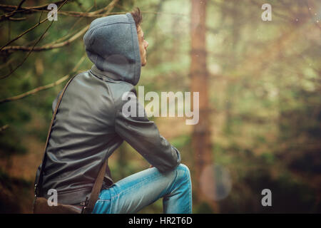 Giovane uomo bello sedersi in una foresta e guardando lontano, vista posteriore Foto Stock