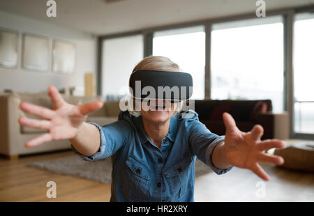 Bella donna senior in denim shirt a casa nel suo salotto indossando la realtà virtuale occhiali, raggiungendo a qualcuno Foto Stock