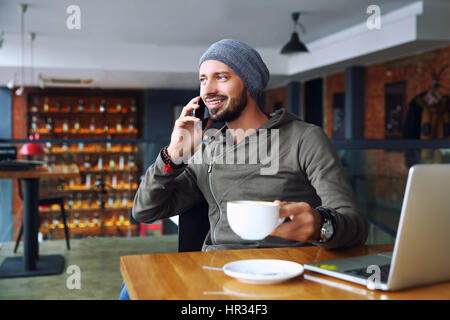 Giovane bello hipster l uomo con la barba in seduta cafe parlando telefono mobile, tenendo tazza di caffè e sorridente. Sul computer portatile sul tavolo di legno. Foto Stock