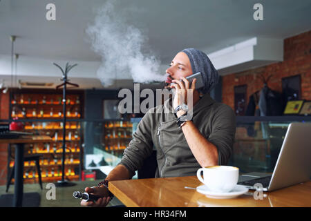 Giovane bello hipster l uomo con la barba seduto nella caffetteria con una tazza di caffè, vaping e rilascia una nube di vapore. Parlando del telefono cellulare e di un po' di pausa. Foto Stock