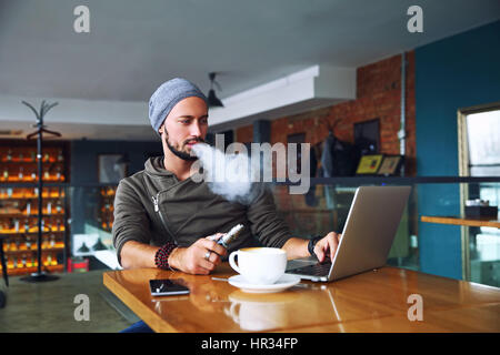 Giovane bello hipster l uomo con la barba seduto nella caffetteria con una tazza di caffè, vaping e rilascia una nube di vapore. Lavorando al computer portatile e di un po' di pausa. Con copia spazio. Foto Stock