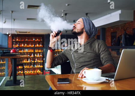 Giovane bello hipster l uomo con la barba seduto nella caffetteria con una tazza di caffè, vaping e rilascia una nube di vapore. Lavorando al computer portatile e di un po' di pausa. Con copia spazio. Foto Stock