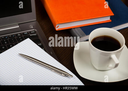 Laptop, tazza di caffè, libri, penna, un foglio di carta e la tazza di caffè in legno scuro, scrivania in ufficio. Foto Stock