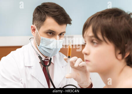 L'uomo medico in maschera medico tenendo la siringa per iniezione al ragazzo paziente Foto Stock