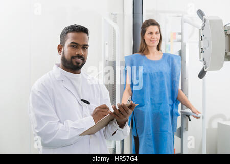 Ritratto del radiologo maschio sulla scrittura di appunti con paziente sottoposto a scansione a raggi x in background in ospedale Foto Stock