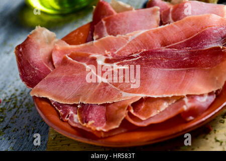 Primo piano di una piastra di terracotta con alcune fette di spagnolo il prosciutto Serrano, il grigio di una tavola in legno rustico e una ampolla con olio di oliva in background Foto Stock