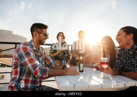 Gruppo multirazziale di amici avente un cocktail party sul tetto. Giovani uomini e donne seduti attorno al tavolo con bevande e ridere. Foto Stock