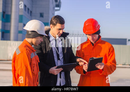 Boss o capo incarica il giovane team di giovani ingegneri con un progetto di costruzione su tablet. Indossano tute e caschi di sicurezza. Azienda moderna Foto Stock