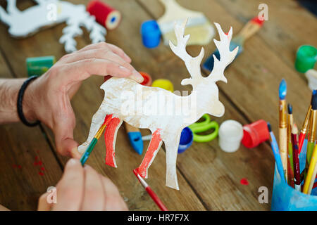 Close-up shot di adulto mani giocattolo di legno cervi e dipinto in rosso, spazzole, tempera, forbici prevista sul tavolo Foto Stock
