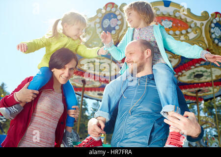 Carefree bambine gioca con ogni altra seduti sulle spalle dei loro sorridente di mezza età i genitori, colorati merry-go-round osservati su BAC Foto Stock