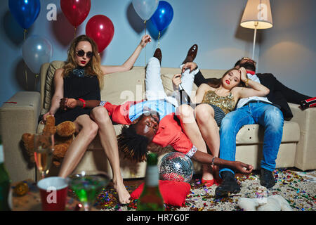 Due belle ragazze rilassante sul divano dopo la festa di Capodanno mentre loro ubriachi amici maschi aventi nap, tutti pavimento coperto di coriandoli Foto Stock