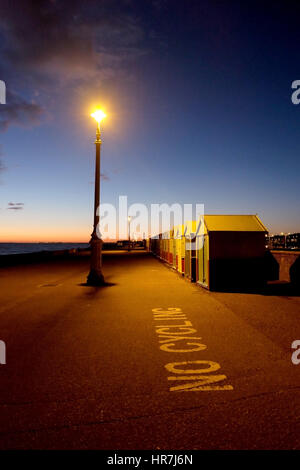 Brighton Seafront, bianco 'n' ciclismo segno scritto sul terreno in primo piano una fila di pittoresca spiaggia di capanne, strada lampada illuminata di notte, cielo blu, Foto Stock
