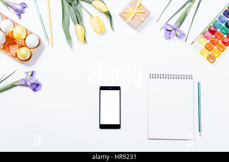 Composizione di Pasqua: fiori, smart phone, dipinto di uova, acquerelli e notebook. Vista dall'alto. Foto Stock