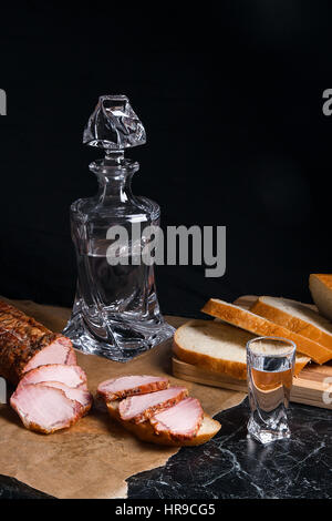 Bottiglia e bicchierini di vodka. Piccolo snack di pane e carne vicino al colpo di vetro. Fette di carne affumicata o prosciutto cotto su marrone carta da imballo. Whea bianco Foto Stock