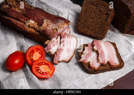 Vista ravvicinata delle fette di pancetta affumicata con segale pane nero su bianco il confezionamento della carta. Fette di pancetta affumicata sul pezzo di pane di segale e di pomodori rossi Foto Stock