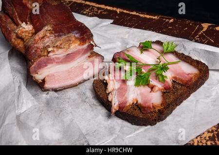 Vista ravvicinata delle fette di pancetta affumicata con segale pane nero su bianco il confezionamento della carta. Naturmort in uno stile rustico su marrone vintage background. Foto Stock