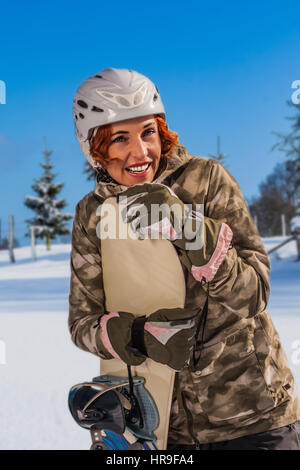 La parte superiore del corpo in aspirazione clima soleggiato di un redheaded, ridendo donna con casco, in piedi sulle piste con lo snowboard in mano. Foto Stock