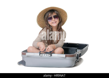 Studio shot di sorridente bambina turistico con cappello di paglia e gli occhiali seduto in valigia. Il tutto è su sfondo bianco. Foto Stock