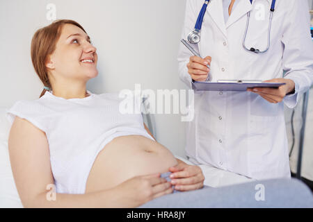 Vista ravvicinata della donna incinta giacente in clinica su check-up, ricevendo buone notizie dal medico e sorridendo allegramente Foto Stock