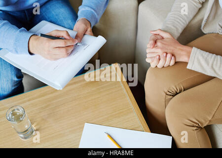 Paziente donna seduta su una poltrona e tenendo le sue mani insieme mentre psicologo maschio il riempimento in medical card, close-up shot Foto Stock