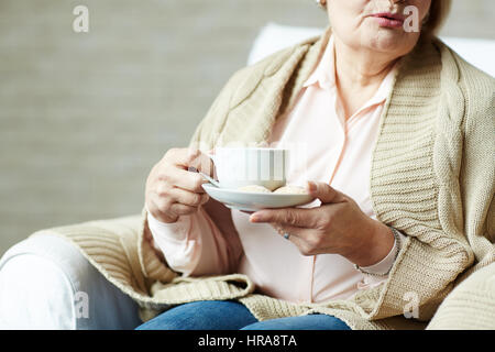 Senior donna in informale in un momento di relax a casa con la tazza di tè e seduto sul divano accogliente Foto Stock