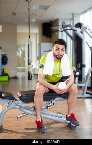 Lavoro in palestra: bello muscoloso uomo seduto sul banco regolabile riposo dopo la pratica con macchine e guardando la telecamera nella sala vuota Foto Stock
