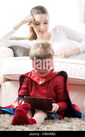 Carino ragazzo supereroe di giocare ai videogiochi seduta sul tappeto con sua madre sullo sfondo. Foto Stock
