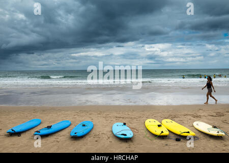 Riga di colore blu e giallo tavole da surf allineate su una spiaggia di fronte ad una calma piatta oceano con una donna camminare davanti al bordo del surf su un cl Foto Stock