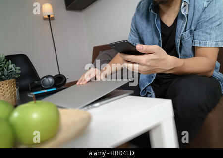 Designer man mano utilizzando compter portatile e i pagamenti mobili shopping online,omni canale,seduto sul divano nel soggiorno,mele verdi nel vassoio in legno Foto Stock