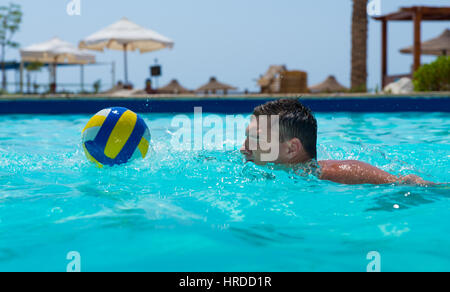 Uomo atletico nuoto e cercando di prendere la palla in una piscina presso l'hotel su una soleggiata giornata estiva Foto Stock