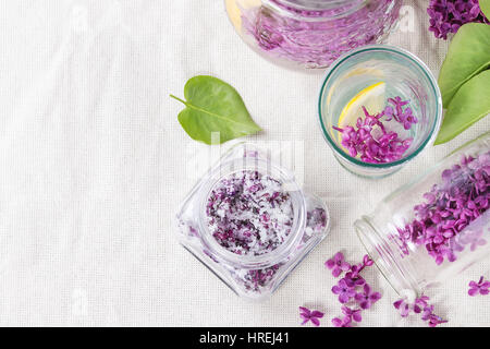 Il vasetto di vetro di lilla fiori in zucchero, il vetro e la brocca di lillà Acqua con limone e il ramo di lilla fresca sulla biancheria da letto bianca tovaglia. Lay piatto Foto Stock