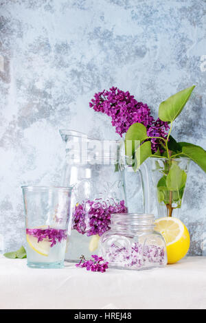 Il vasetto di vetro di lilla fiori in zucchero, il vetro e la brocca di lillà Acqua con limone e il ramo di lilla fresca sulla biancheria da letto bianca tovaglia con textur blu Foto Stock