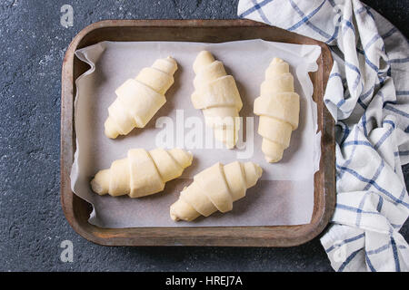 Materie crudi croissant sulla carta da forno nel vecchio forno il vassoio con carta asciugatutto per black texture di sfondo di calcestruzzo. Vista superiore Foto Stock