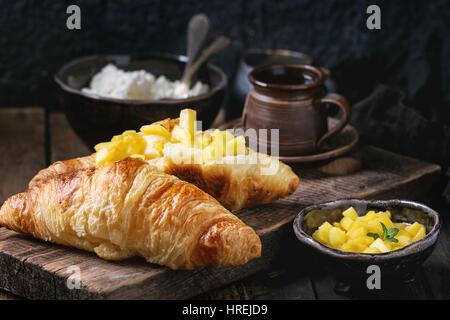 La prima colazione con due cornetti, burro, formaggio, caffè e fette di mango fruit, servita su legno tagliere sopra il vecchio sfondo di legno. Chiudere u Foto Stock