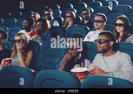 Gruppo di africani e caucasici guardando film in 3D bicchieri nella moderna sala cinema. Giovani mangiare popcorn e beve coca cola, trascorrendo del tempo libero Foto Stock