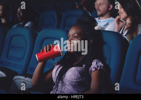 Giovane ragazza africana di bere cola e guardare film. Bella donna elegante con il taglio di capelli seduto in una sedia confortevole in cinema e seriamente guardando a Foto Stock