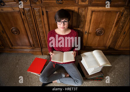 Giovane studentessa seduto sul pavimento con aprire il Blocco note e matita e con tanti libri intorno, guardando la visualizzazione della telecamera dalla parte superiore Foto Stock