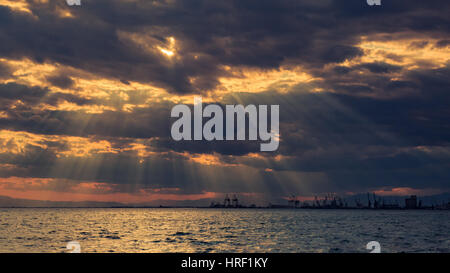 Luminoso nuvoloso tramonto sul mare che si affaccia su sagome delle gru nella parte industriale della città in Grecia. Foto Stock