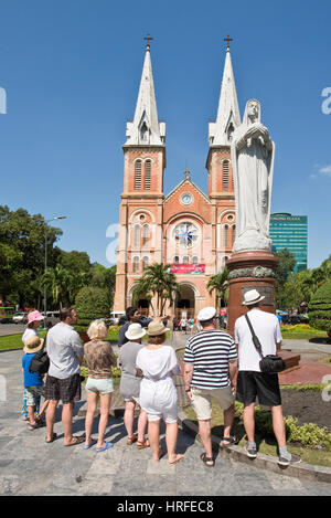 La cattedrale di Notre Dame Basilica a Ho Chi Minh City con i turisti e un tour guida accanto a "Nostra Signora della Pace' statua in una giornata di sole con cielo blu. Foto Stock