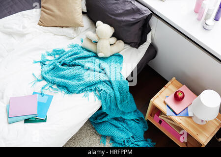 Letto disfatto di una ragazza con il bianco lonely orsacchiotto e coperta di blu che pendono sul pavimento, ad alto angolo di visione Foto Stock