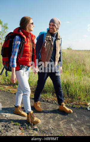 Coppia di giovani viaggiatori con zaini sorridente e parlare mentre si cammina insieme su escursioni via tenendo le mani sotto la luce diretta del sole Foto Stock