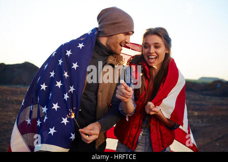 Ritratto di giovane coppia felice azienda botti, ridendo e entrambi avvolti nella bandiera americana al tramonto Foto Stock