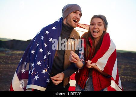 Ritratto di giovane coppia felice ridendo e azienda botti, entrambi avvolti nella bandiera americana al tramonto in montagna Foto Stock