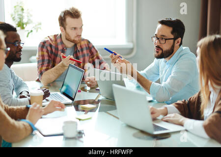 Multi-etnico team di progettisti ambiziosi in informale suggerendo idee riguardanti il loro nuovo progetto seduti insieme in sala riunioni Foto Stock