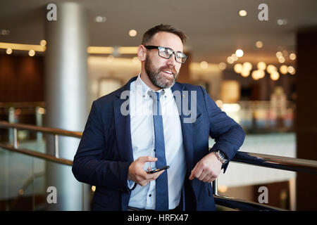 Ritratto di barbuto imprenditore maturo indossando elegante abito formale e bicchieri appoggiata sulla ringhiera in ufficio edificio tenendo lo smartphone a portata di mano Foto Stock