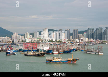 Una vista del porto di Sanya in Cina con barche da pesca e le navi ormeggiate ed i grattacieli della città dietro. Foto Stock