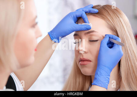 Vista laterale del cosmetologo in guanti spiumatura sopracciglia per la bella donna che utilizza il forcipe. Donna con gli occhi chiusi godendo di procedura di rendere perma Foto Stock