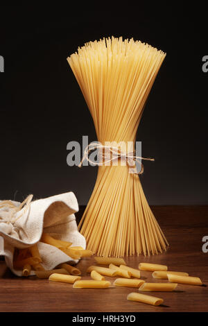 Asciugare gli spaghetti legato con corda e le penne rigate pasta cadere al di fuori della sacca di tela su legno scuro tabella su sfondo grigio Foto Stock